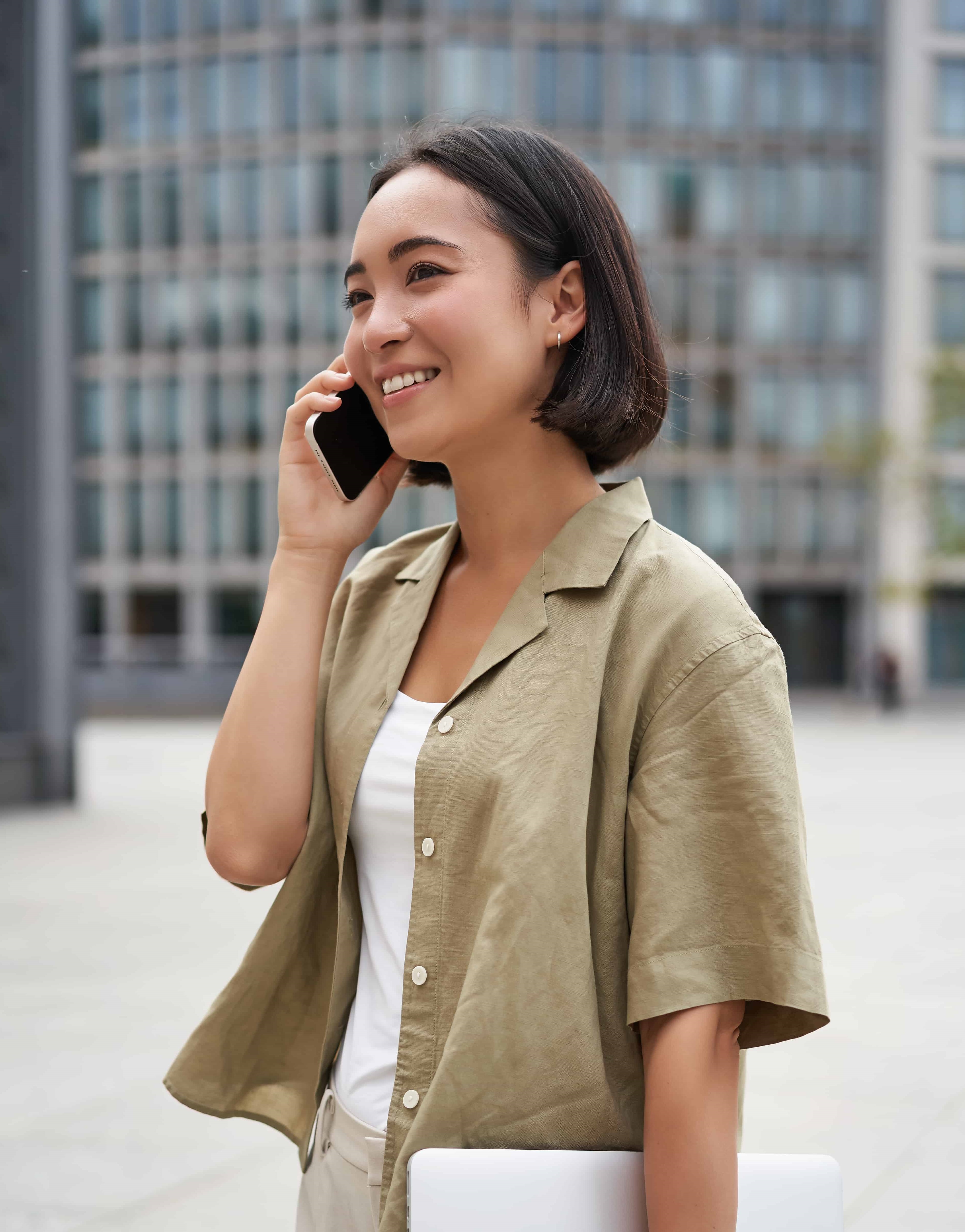 vertical shot young asian digital nomad girl talks mobile phone walks street with laptop